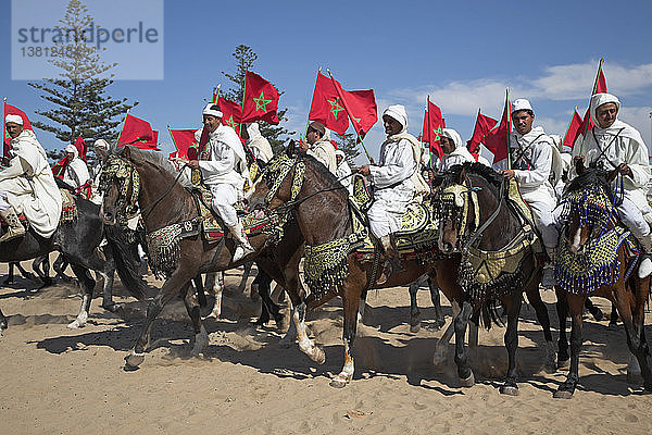Männer auf Pferden in traditionellen weißen Gewändern schwenken die rote Nationalflagge  Essaouira  Marokko