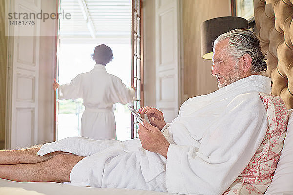 Älterer Mann im Bademantel mit digitalem Tablet auf einem Hotelbett