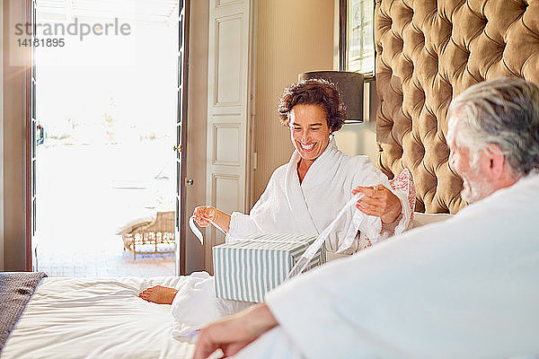 Ehemann beobachtet glückliche Frau beim Öffnen des Geschenks auf dem Hotelbett