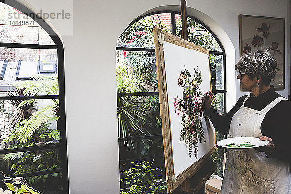 Ältere Frau mit Brille  rotem Kleid und weißer Schürze steht im Atelier und arbeitet an der Malerei von rosa Teerosen  Blättern  Beeren und anderen Blumen.