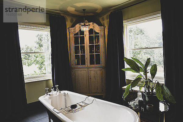 Innenansicht des Badezimmers mit Eckschrank aus Holz zwischen den Schiebefenstern  Badewanne mit Rollladen aus Messing.