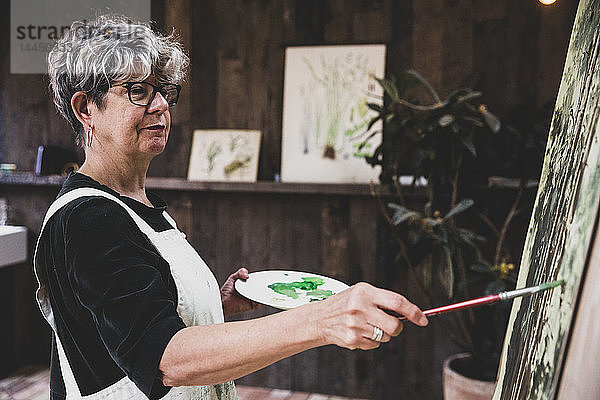 Ältere Frau mit Brille  schwarzem Oberteil und weißer Schürze steht im Atelier und arbeitet an der Bemalung von Bäumen im Wald.