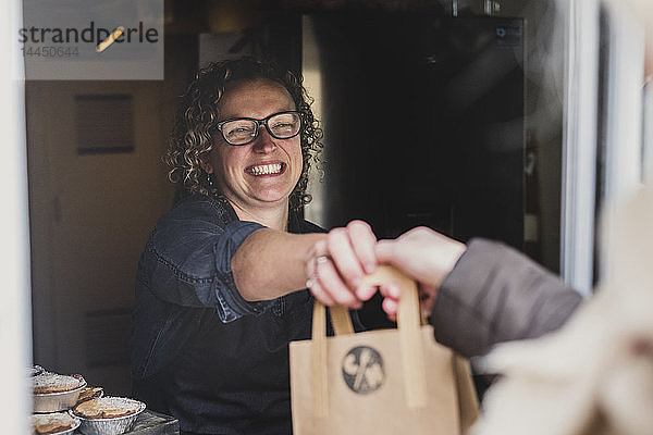 Lächelnde Frau mit Brille  die eine braune Einkaufstasche aus Papier durch das Fenster einer Bäckerei reicht.