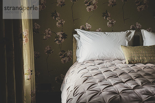 Innenansicht des Schlafzimmers mit Vorhängen und Tapeten mit Blumenmuster  zartrosa Bettdecke und weißen Kissen auf dem Doppelbett.