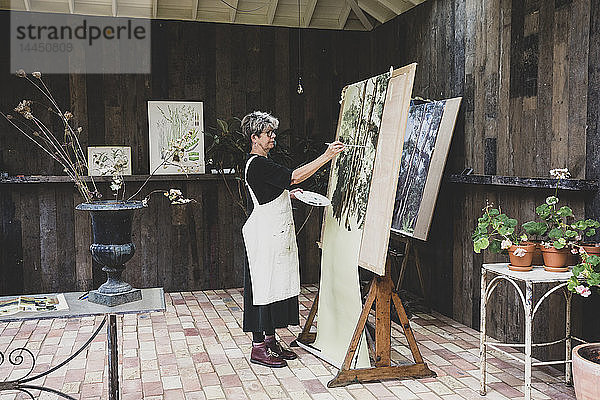 Ältere Frau mit Brille  schwarzem Oberteil und weißer Schürze steht im Atelier und arbeitet an der Bemalung von Bäumen im Wald.