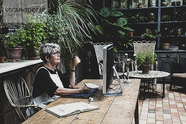 Ältere Frau mit Brille  schwarzem Oberteil und weißer Schürze sitzt an einem Holztisch und arbeitet am Desktop-Computer.