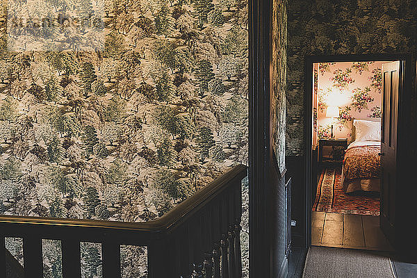 Innenansicht des Treppenabsatzes mit Holzgeländer  Tapete mit grünem und braunem Baummuster  offene Tür zum Schlafzimmer mit Holzfußboden  Doppelbett und Blumentapete.