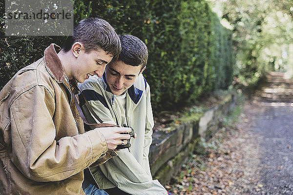 Ein junger Mann und ein Teenager mit kurzen braunen Haaren in Freizeitjacken sitzen auf einer eingezäunten Steinmauer und schauen auf das Bild einer Digitalkamera.