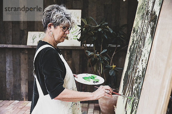 Ältere Frau mit Brille  schwarzem Oberteil und weißer Schürze steht im Atelier und arbeitet an der Bemalung von Bäumen im Wald.