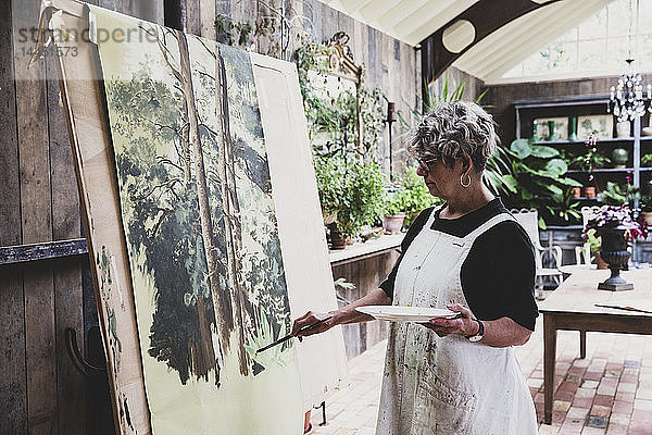 Ältere Frau mit Brille  schwarzem Oberteil und weißer Schürze steht im Atelier und arbeitet an der Bemalung von Bäumen im Wald.