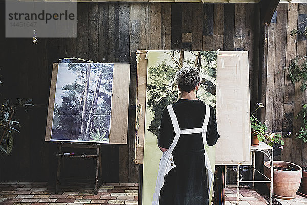 Ältere Frau in schwarzem Kleid und weißer Schürze steht im Atelier und arbeitet an der Bemalung von Bäumen im Wald.