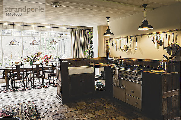 Innenansicht der Küche mit Steinfliesenboden  Herd und Butlerspüle  offener Grundriss zum Wintergarten mit antikem Holztisch und Stühlen.