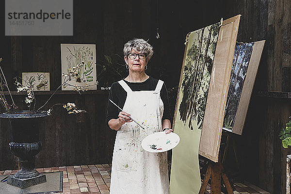 Ältere Frau mit Brille  schwarzem Oberteil und weißer Schürze steht im Atelier und arbeitet an der Bemalung von Bäumen im Wald.