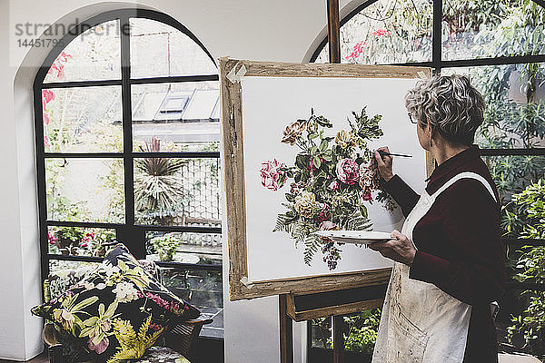 Ältere Frau mit Brille  rotem Kleid und weißer Schürze steht im Atelier und arbeitet an der Malerei von rosa Teerosen  Blättern  Beeren und anderen Blumen.