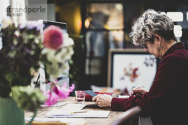 Ältere Frau mit Brille und rotem Kleid sitzt an einem Holztisch und schaut auf ein digitales Tablett.
