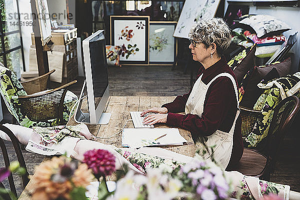 Ältere Frau mit Brille  rotem Kleid und weißer Schürze sitzt an einem Holztisch in einem Studio und arbeitet am Desktop-Computer.