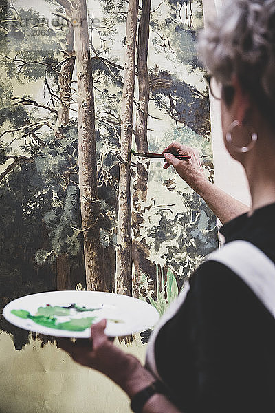 Ältere Frau mit Brille  schwarzem Oberteil und weißer Schürze steht im Atelier und arbeitet an der Bemalung von Bäumen im Wald.