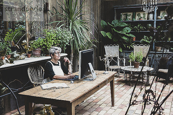 Ältere Frau mit Brille  schwarzem Oberteil und weißer Schürze sitzt an einem Holztisch und arbeitet am Desktop-Computer.