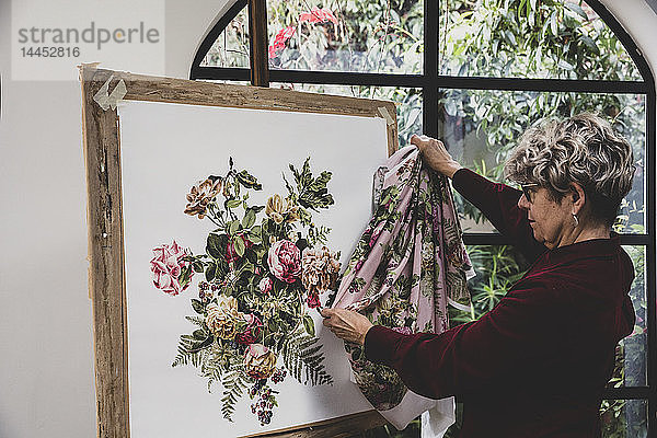 Eine ältere Frau mit Brille und rotem Kleid steht im Atelier und vergleicht Stoffmuster mit einem Gemälde aus rosa Teerosen  Blättern  Beeren und anderen Blumen.