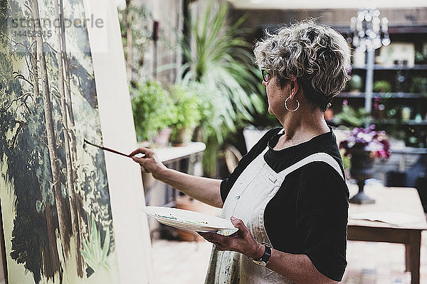 Ältere Frau mit Brille  schwarzem Oberteil und weißer Schürze steht im Atelier und arbeitet an der Bemalung von Bäumen im Wald.