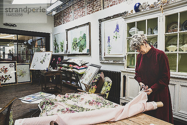 Ältere Frau mit Brille und rotem Kleid steht im Atelier und schneidet mit einer Schere rosa Stoff mit Blumenmuster zurecht.
