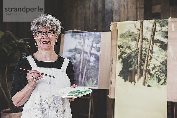 Ältere Frau mit Brille  schwarzem Oberteil und weißer Schürze steht im Atelier und arbeitet an der Bemalung von Bäumen im Wald.