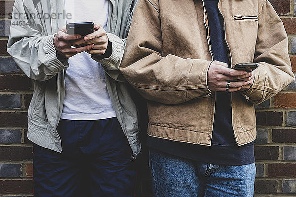 In der Mitte zwei junge Männer in Freizeitjacken  die nebeneinander an einer Ziegelmauer stehen und mit ihren Mobiltelefonen SMS schreiben.