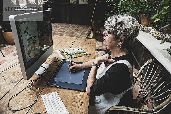 Ältere Frau mit Brille  schwarzem Oberteil und weißer Schürze sitzt an einem Holztisch und arbeitet am Desktop-Computer.