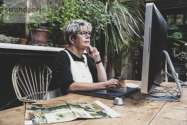 Ältere Frau mit Brille  schwarzem Oberteil und weißer Schürze sitzt an einem Holztisch und arbeitet am Desktop-Computer.