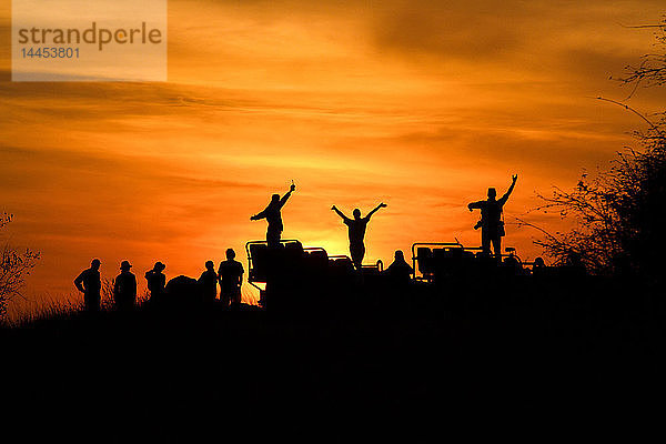 Eine Silhouette von Menschen und einem Fahrzeug  Menschen mit Händen in der Luft  vor Sonnenuntergang  orangefarbenem und gelbem Himmel
