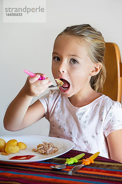 Ein kleines Mädchen  das am Tisch sitzend Kartoffeln und Huhn isst.