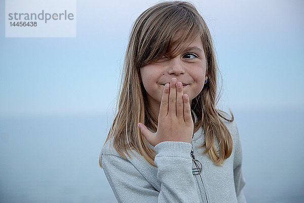 Porträt eines ausdrucksstarken kleinen Mädchens mit Hand vor dem Mund vor dem Meer bei Einbruch der Dunkelheit.