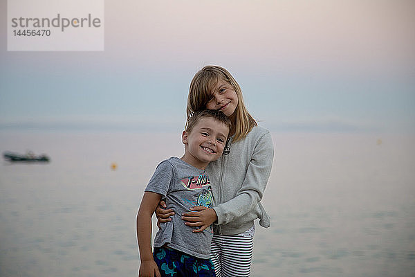 Ein kleines Mädchen mit ihrem Bruder auf dem Arm posiert zärtlich vor dem Meer bei Einbruch der Dunkelheit.