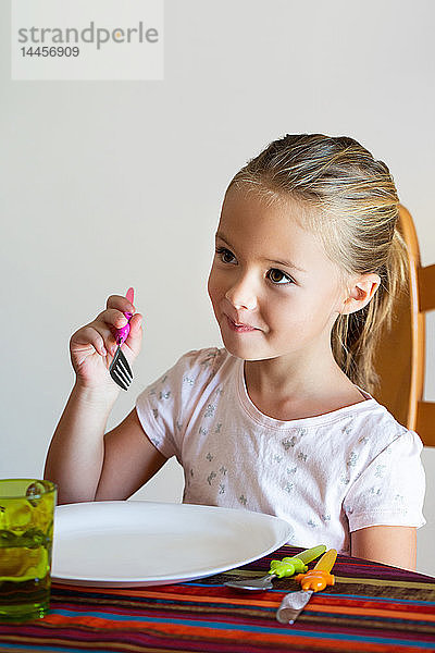 Ein hübsches  lächelndes kleines Mädchen  das an einem Tisch vor einem leeren weißen Teller sitzt und seine Gabel hält.