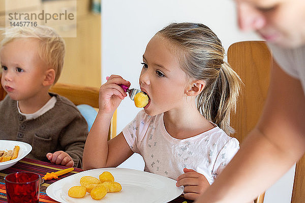 Ein kleines Mädchen  das mit seinem kleinen Bruder Kartoffeln isst  sitzt am Tisch  während die Mutter sie ködert.