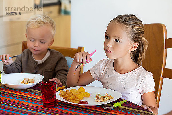 Ein kleines Mädchen isst Kartoffeln und Hühnchen mit ihrem kleinen Bruder am Tisch.
