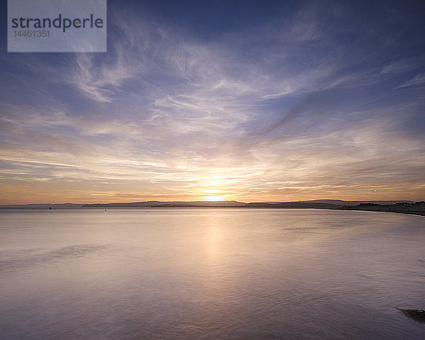 Mit interessanten Wolkenfetzen geht die Sonne über dem Wasser von Exmouth  Devon  England  Vereinigtes Königreich  unter.