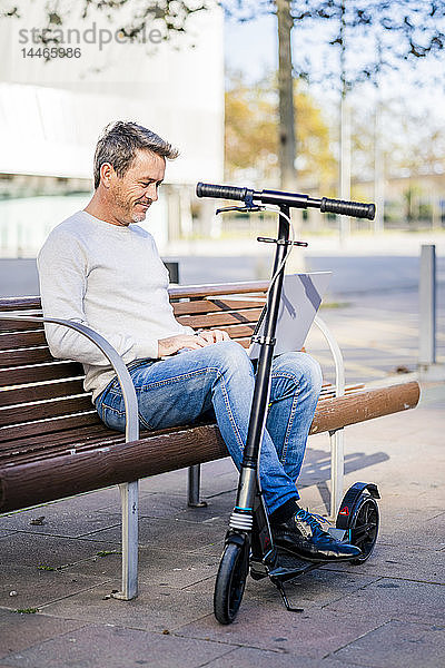 Lässiger Geschäftsmann mit Kickroller  der auf einer Bank sitzt und entspannt in der Stadt arbeitet