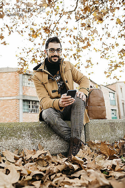 Spanien  Igualada  Porträt eines lächelnden Mannes mit Handy  der in der herbstlichen Stadt sitzt