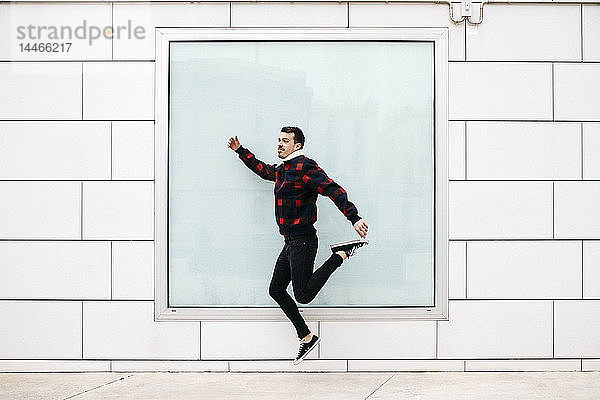 Junger Mann in legerer Kleidung springt mit einer weißen Wand und einem großen Fenster im Hintergrund