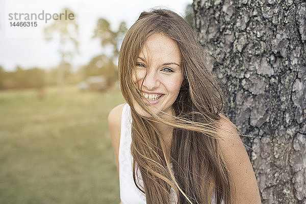 Porträt einer lachenden jungen Frau  die sich an einen Baumstamm lehnt