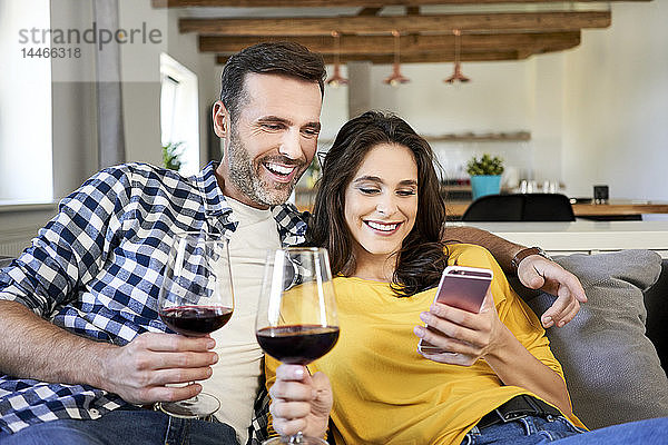 Ehepaar sitzt im Wohnzimmer  trinkt Rotwein und benutzt ein Smartphone