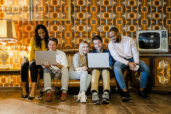 Gruppe von lächelnden Menschen  die auf einer Couch im alten Wohnzimmer sitzen und sich Laptops teilen