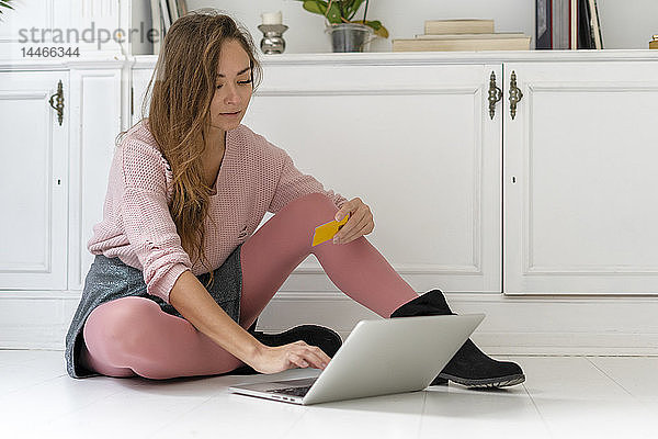 Junge Frau sitzt auf dem Boden und macht eine Online-Zahlung mit ihrer Kreditkarte