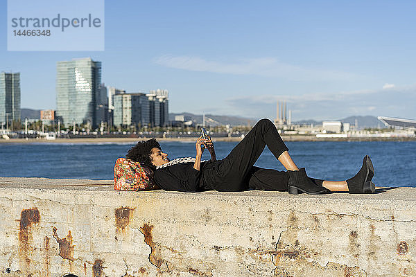 Spanien  Barcelona  schwarz gekleidete Frau  die mit einem Smartphone an einer Wand vor dem Meer liegt