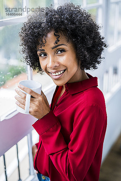 Porträt einer lächelnden Frau mit Tasse Kaffee am Fenster zu Hause