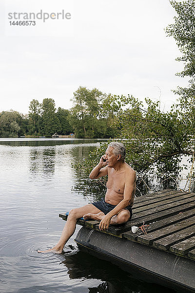 Älterer Mann sitzt auf einem Steg an einem See und telefoniert