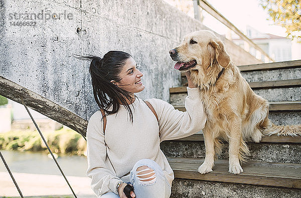 Lächelnde junge Frau streichelt ihren Golden-Retriever-Hund auf einer Treppe im Freien