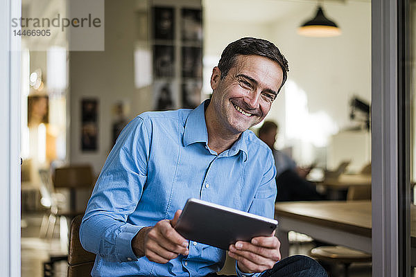 Lächelnder Geschäftsmann mit Tablette sitzt an offener französischer Tür