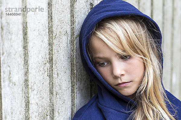 Bildnis eines traurigen blonden Mädchens mit blauer Kapuzenjacke  das an einer Holzwand lehnt